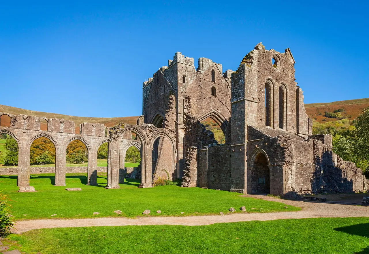 Ruins of a medieval stone abbey or castle with arched columns and towers set in a grassy valley under clear blue sky.