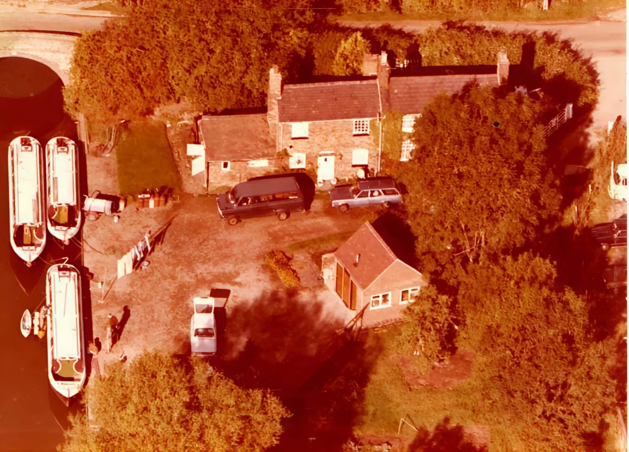Aerial view of a riverside mooring with narrowboats docked alongside brick buildings and parked vehicles.