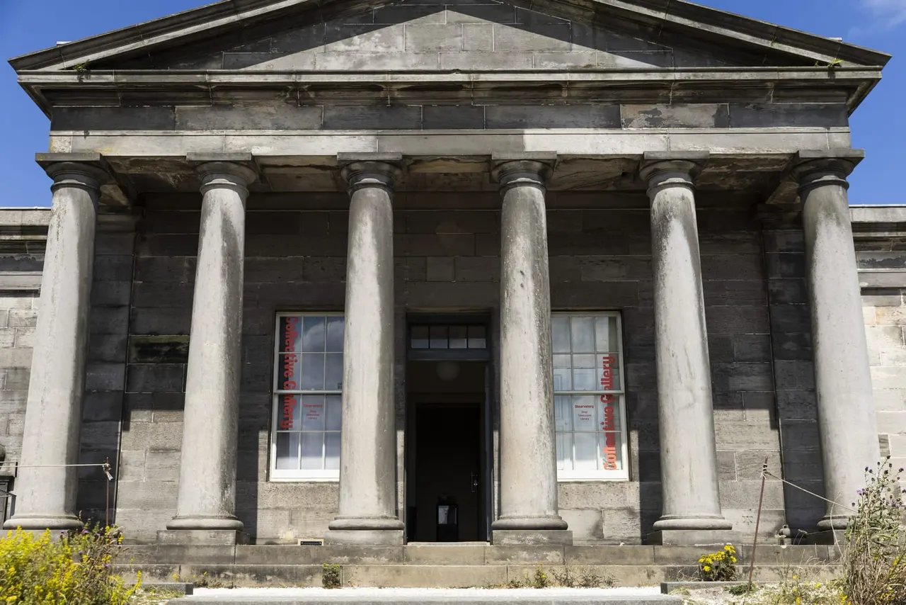 Classical stone building with columned front entrance and triangular pediment, clear blue sky above.