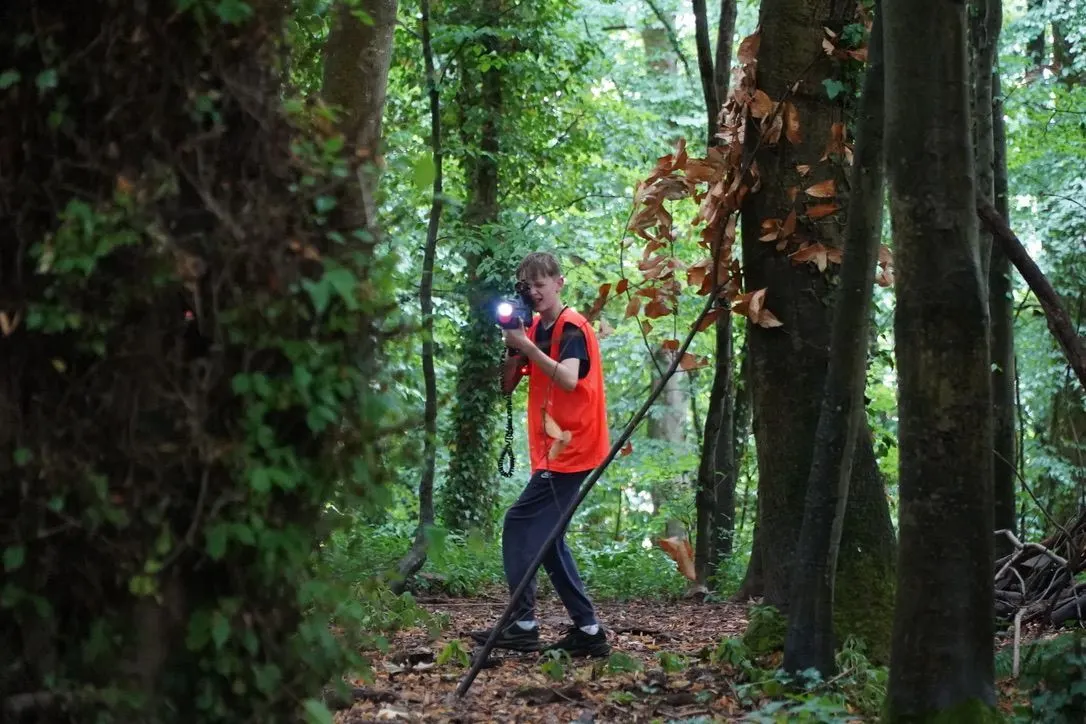 Child in red shirt holding a torch whilst walking through a dense woodland path surrounded by tall trees.