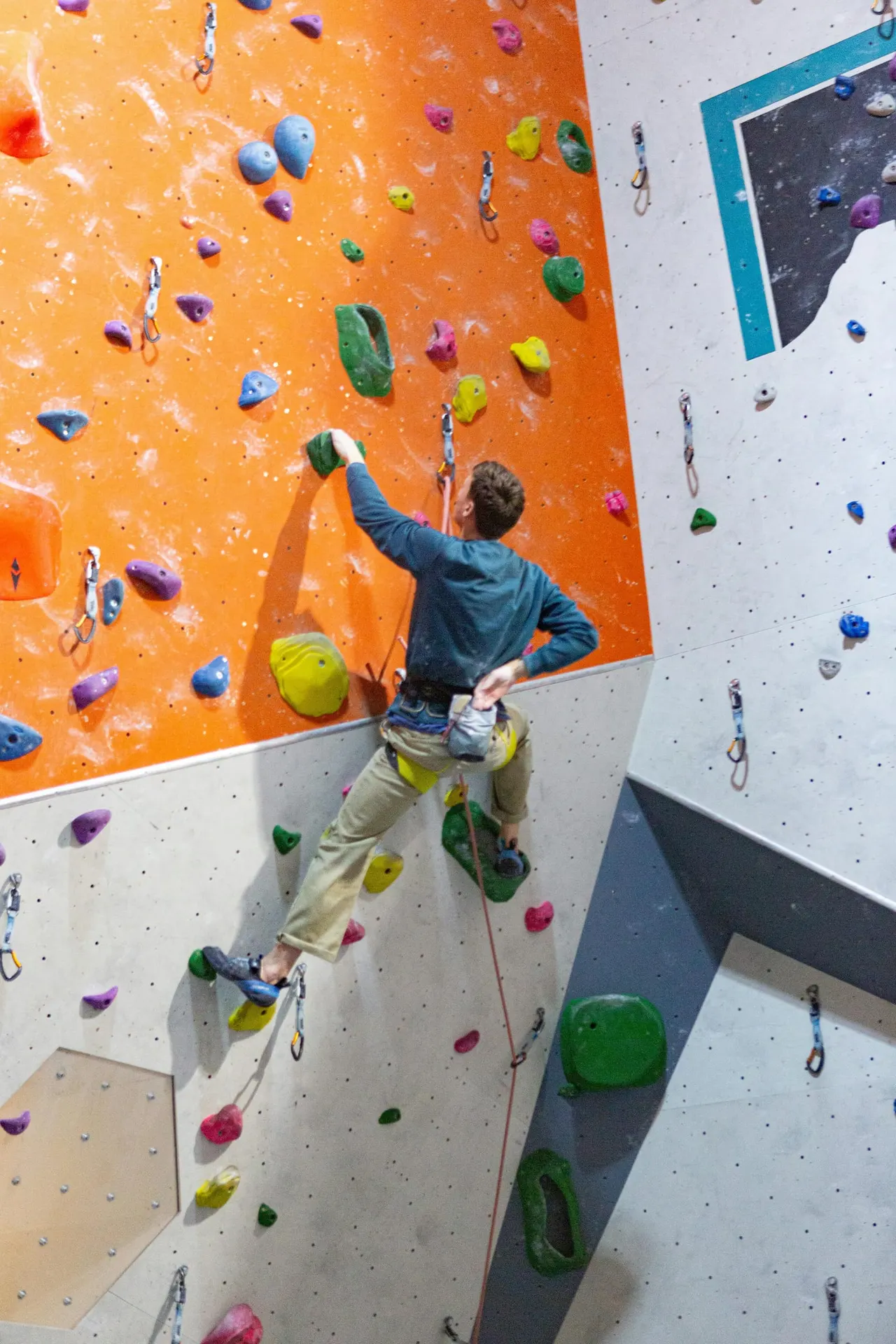 Child in helmet rock climbing an indoor climbing wall with coloured handholds, secured with a rope.