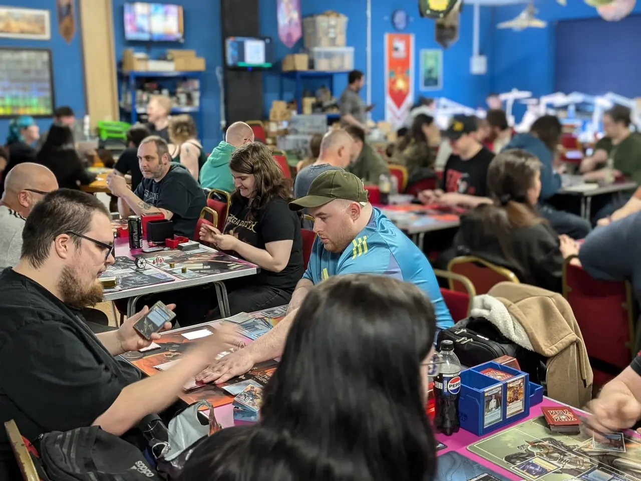 Large group of people playing trading card games at tables in a bright blue indoor gaming venue.
