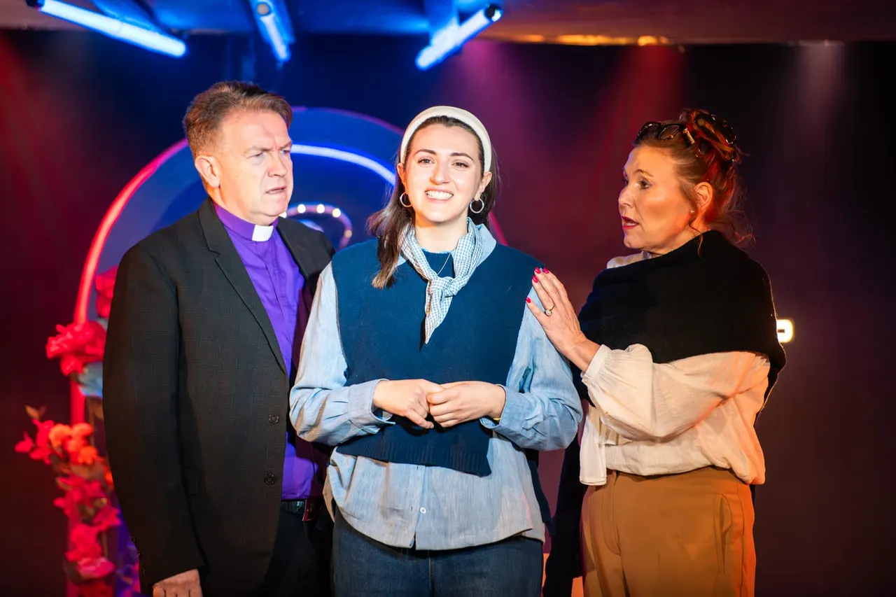Three actors perform a scene on stage under blue and red neon lighting, with colourful stage set behind them.