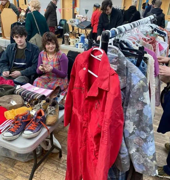 People browsing second-hand clothing and shoes at an indoor market or car boot sale event.