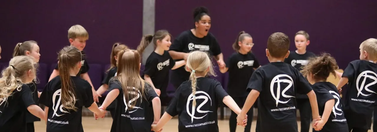 A group of children in black Razzamataz t-shirts perform together on stage against a purple backdrop.