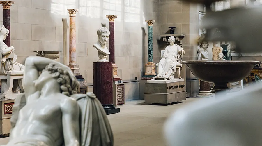 Classical sculpture gallery with marble busts and statues displayed on ornamental pedestals and columns.