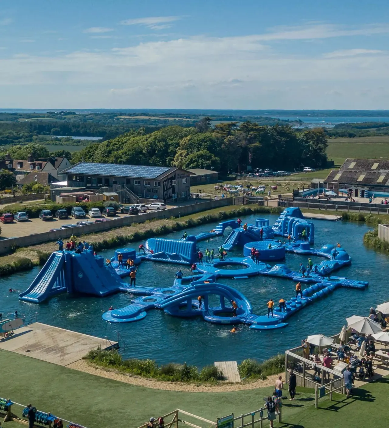 Visitors enjoying a large blue inflatable aqua park with slides and obstacles in a water channel, surrounded by landscaped grounds and countryside views.