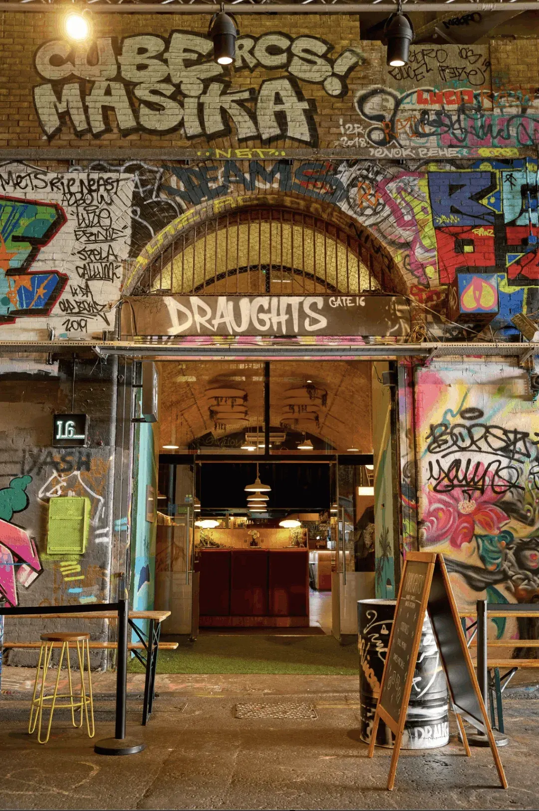 Street-level storefront entrance to a bar with colourful graffiti-covered brick walls and an ornate arched window above.
