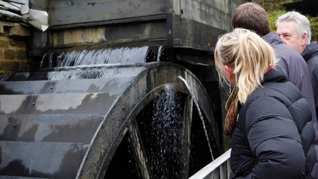 Visitors observe water flowing over a large wooden waterwheel at an industrial heritage site.