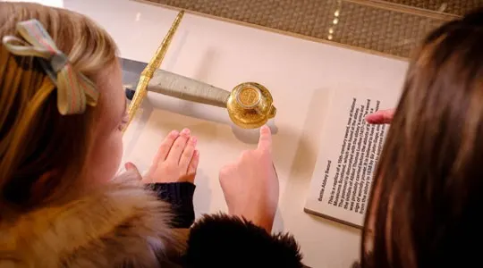 Two people in period costume examine a golden spherical object on display behind glass.