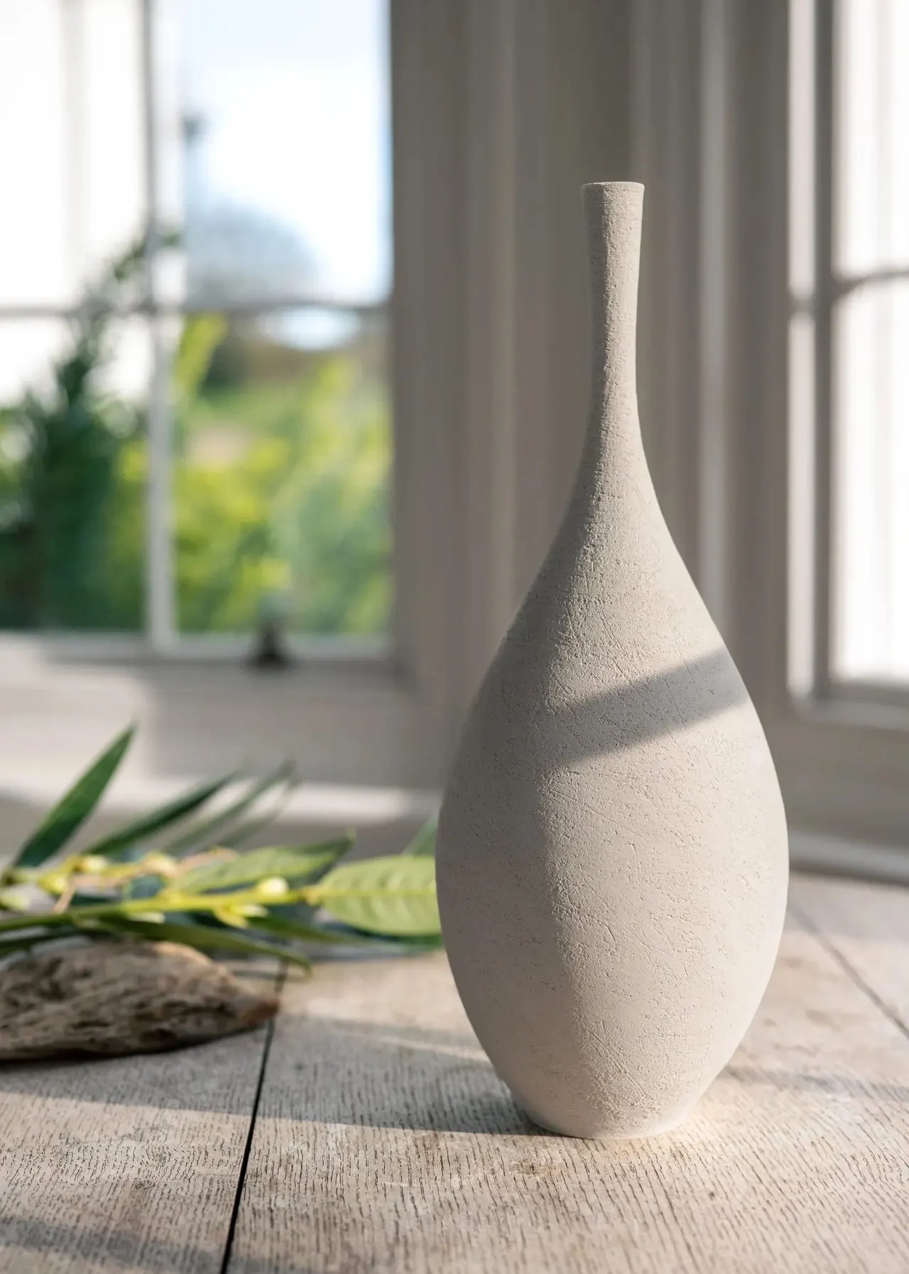 A cream-coloured ceramic bottle vase sits on wooden flooring beside greenery, with windows and curtains behind.