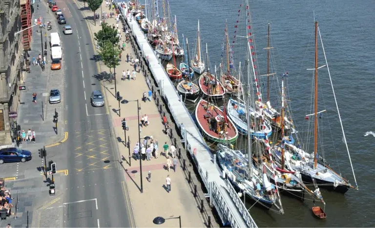 Tall ships with red and white hulls moored at a waterfront promenade beside a busy city street.
