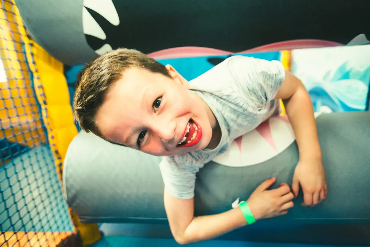 Young child smiling at camera whilst playing in an indoor soft play area with inflatable structures.
