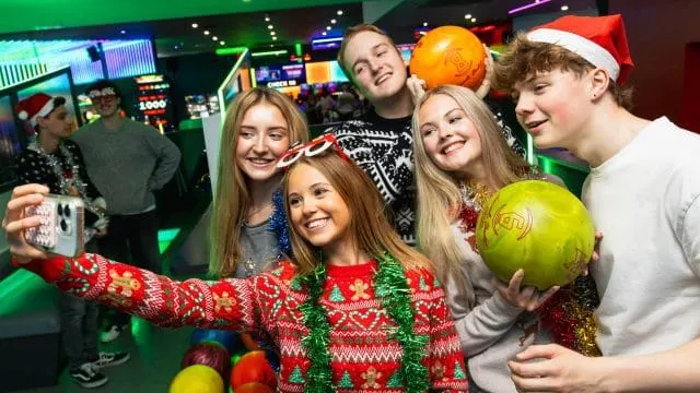 Group of young people holding bowling balls and taking a selfie in a festive bowling alley during Christmas.