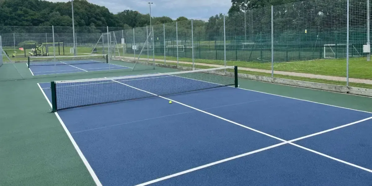 Multiple pickleball courts with blue and green playing surfaces, nets, and fencing, with trees and sports equipment visible in the background.