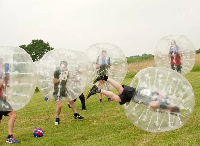 People playing bubble football in a grassy field, enclosed in large inflatable transparent spheres.