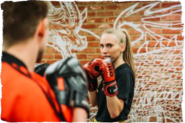 Woman wearing red boxing gloves sparring with male instructor in black and orange sportswear against brick wall.