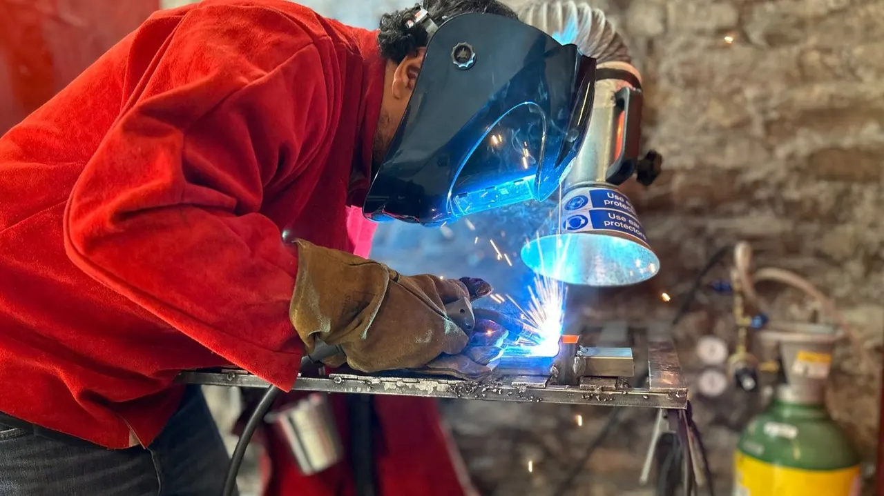 Person in red jacket and welding helmet arc welding metal on a workbench in an industrial workshop.