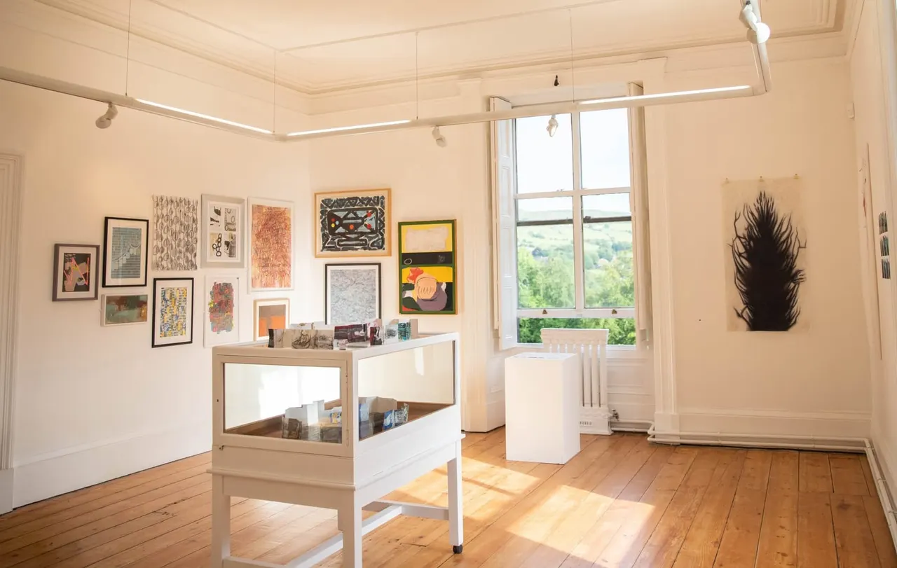 Interior of an art gallery with framed artwork on cream walls, display cases, wooden flooring, and large windows overlooking green countryside.