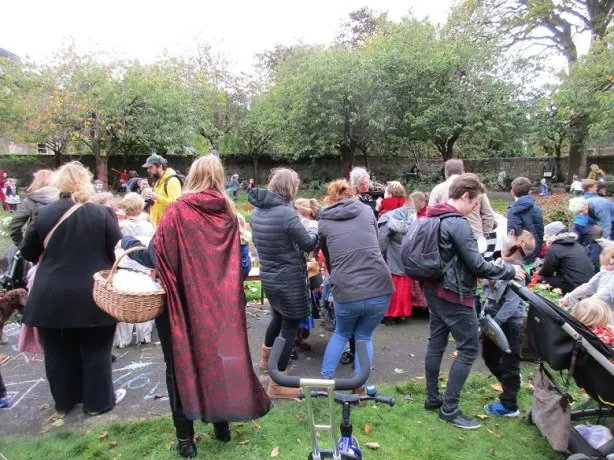 A crowd of people gathered outdoors in a park surrounded by mature trees, with some holding baskets and picnic items.