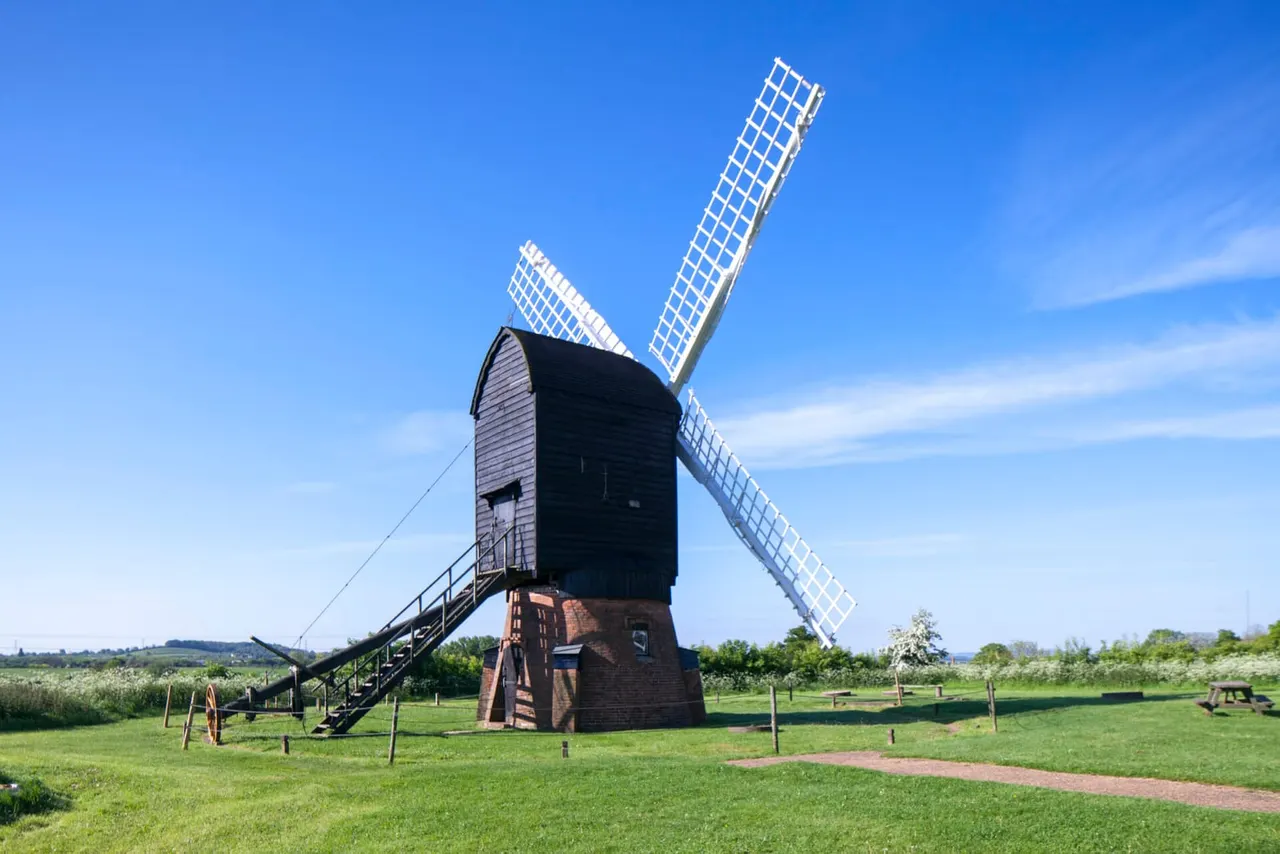 A restored brick windmill with white sails stands in a green field under blue sky.