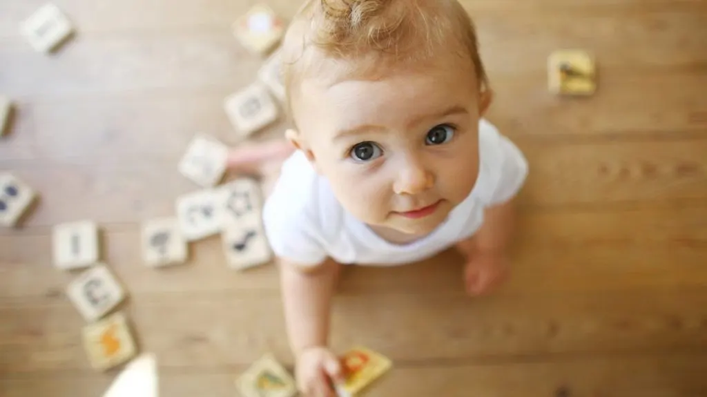 Toddler in white shirt looking up at camera surrounded by letter blocks on wooden floor.
