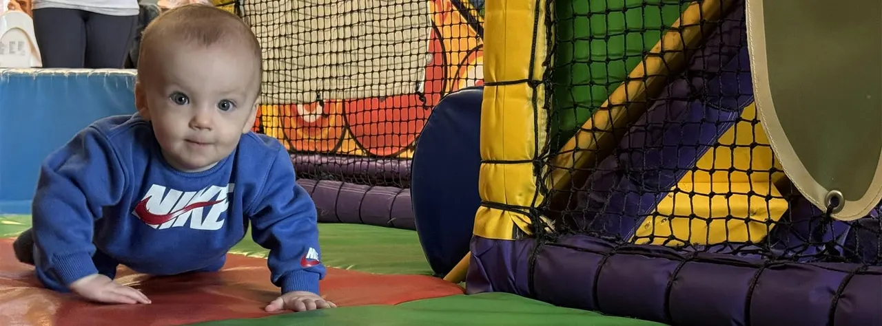 Toddler in blue sweatshirt crawling through a colourful indoor play structure with netting and padded elements.