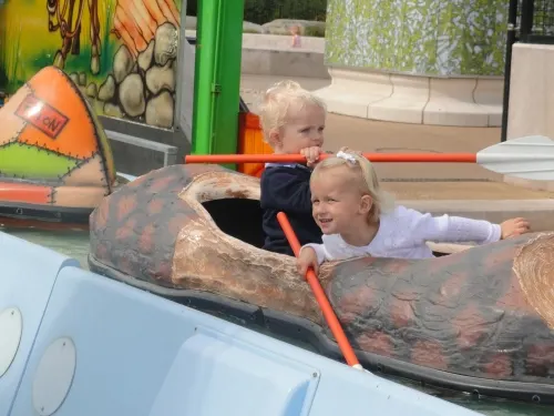 Two young children sit in a log-shaped ride vehicle, holding orange poles and smiling at a leisure park.