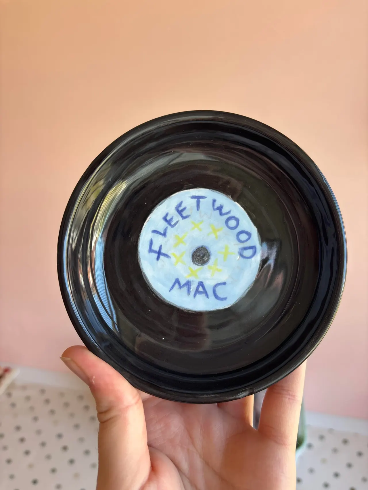 A hand holding a black vinyl record with a handwritten white label reading "Fleetwood Mac" against a pale pink background.
