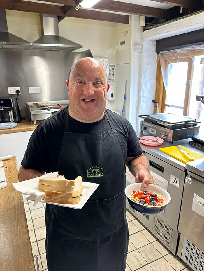 A man in an apron smiles whilst holding a bowl of colourful breakfast cereal and slices of toast in a kitchen.