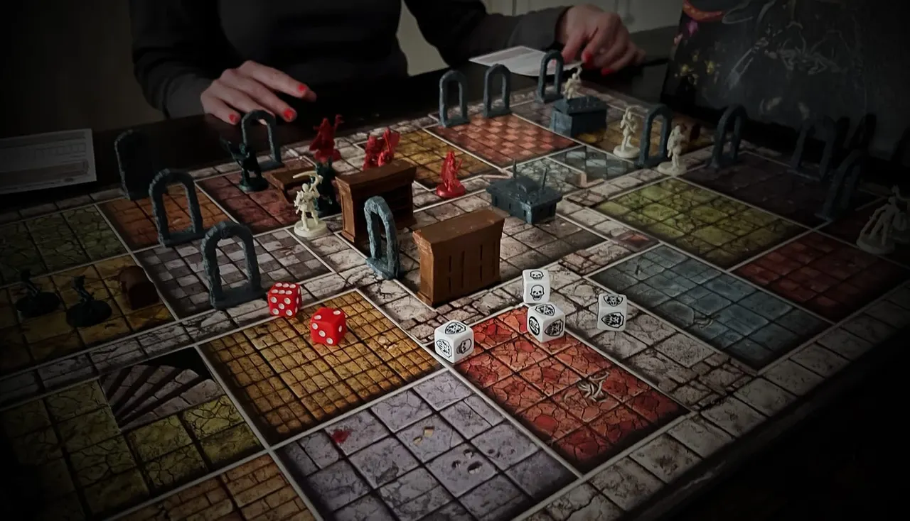 Players' hands above a dungeon-crawler board game with miniature figures, dice, and interlocking game tiles.