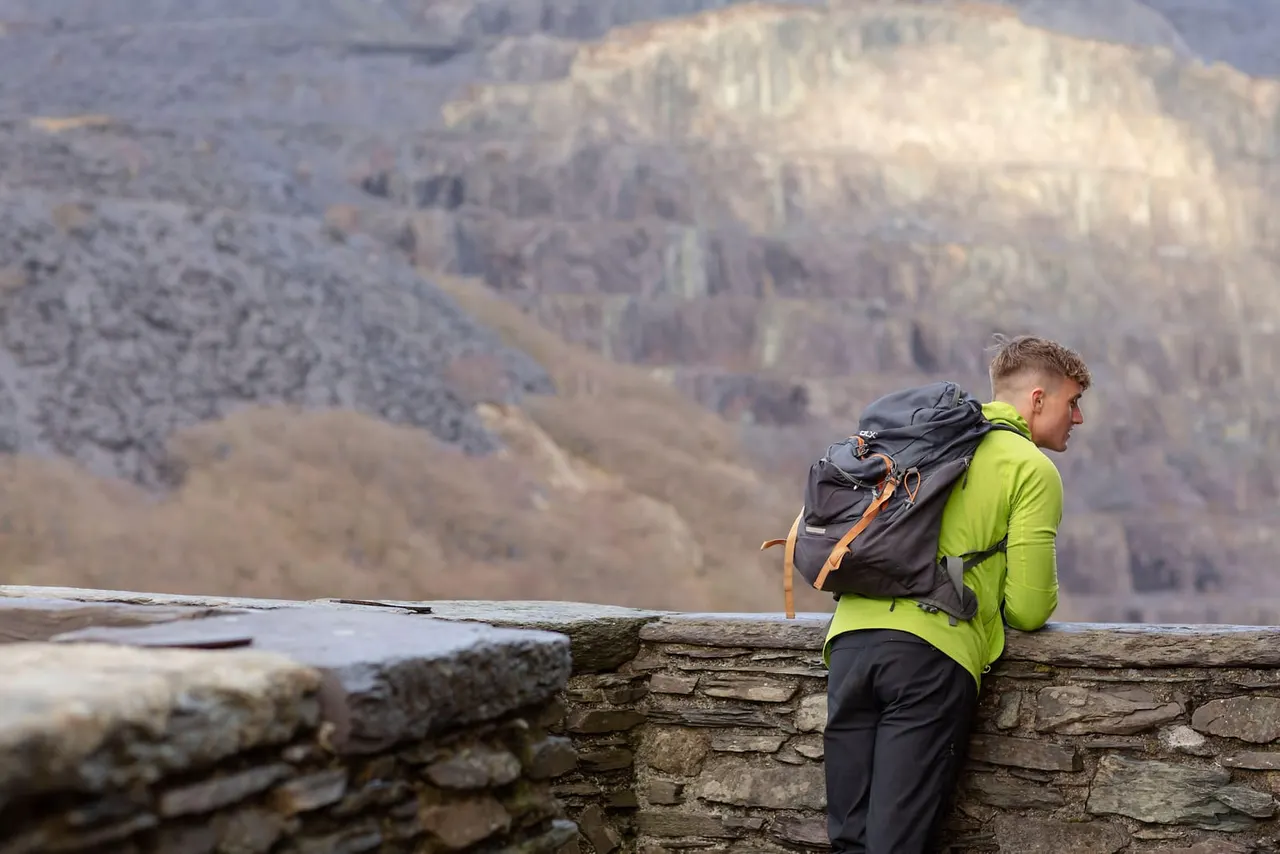 A hiker in a bright yellow jacket and backpack stands on a stone wall overlooking a forested valley.