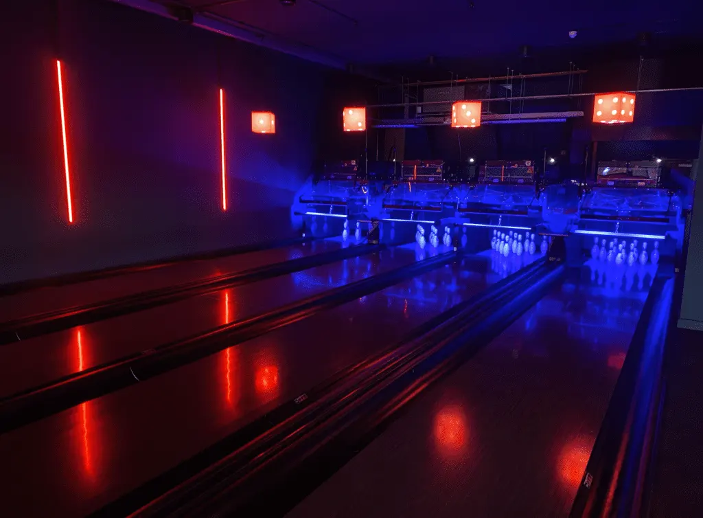 Indoor bowling alley with multiple lanes illuminated by red and blue neon lighting and overhead scoring displays.