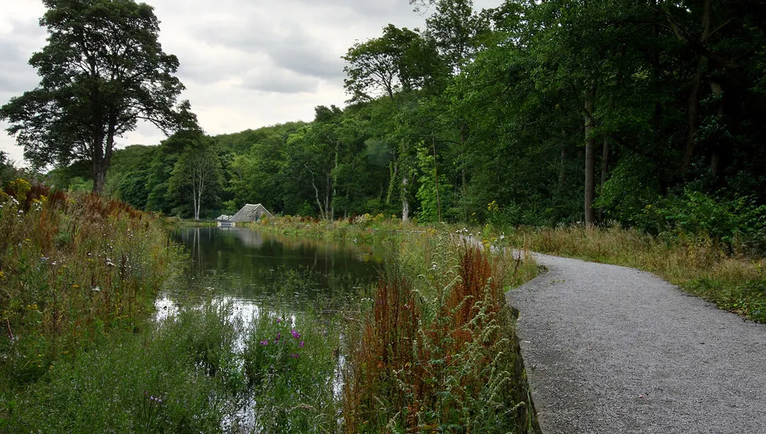 Gravel path beside a canal with wildflowers and trees, a boathouse visible across the water.