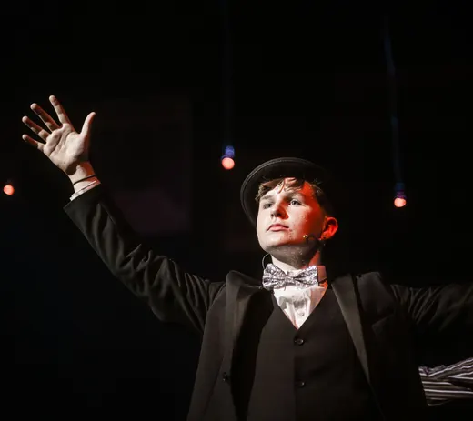 Performer in formal attire with hat and bow tie gestures dramatically on stage under spotlights.