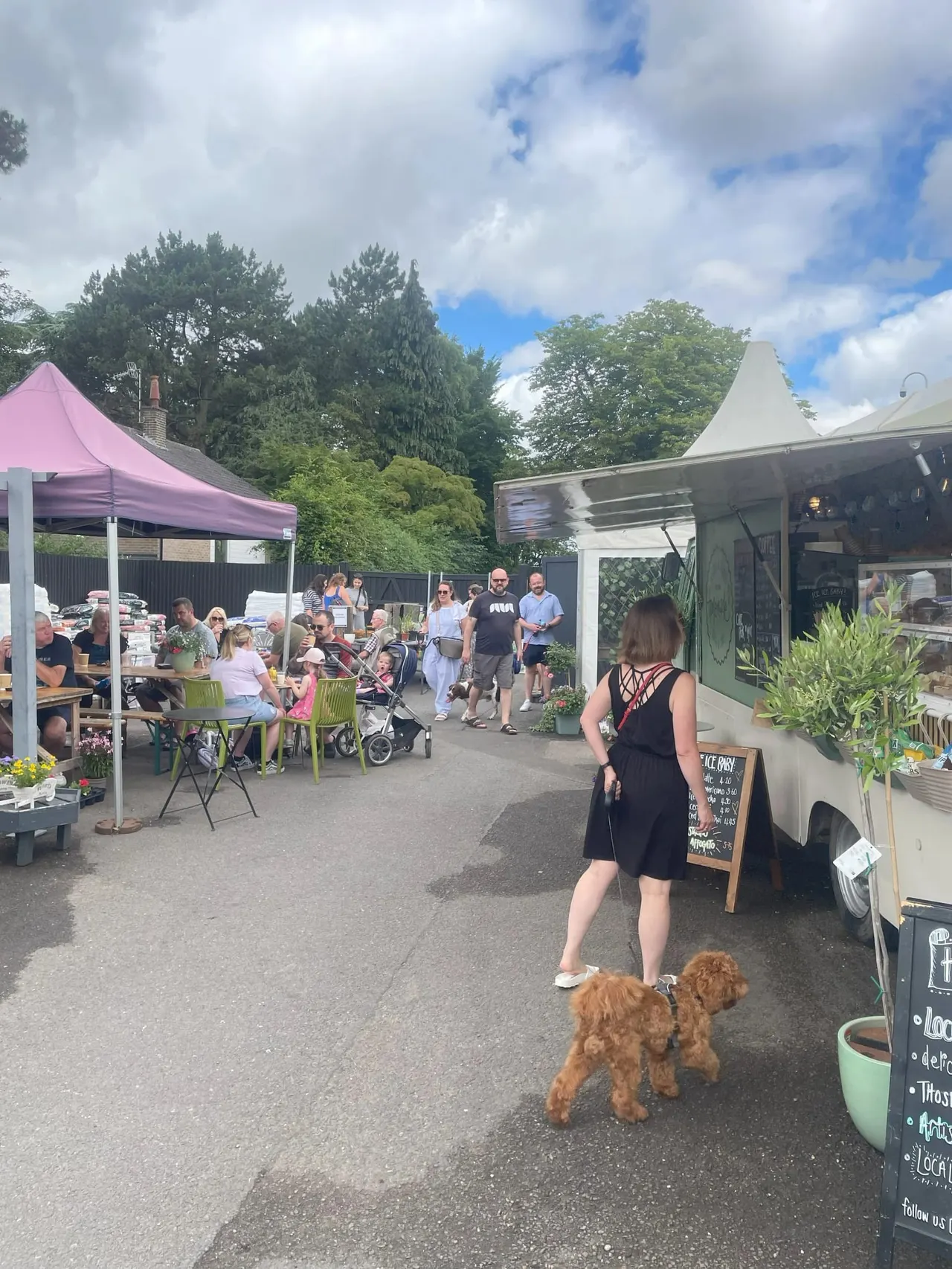 Woman with a small reddish dog walks past food trucks and pink marquee at an outdoor cafe event with visitors.