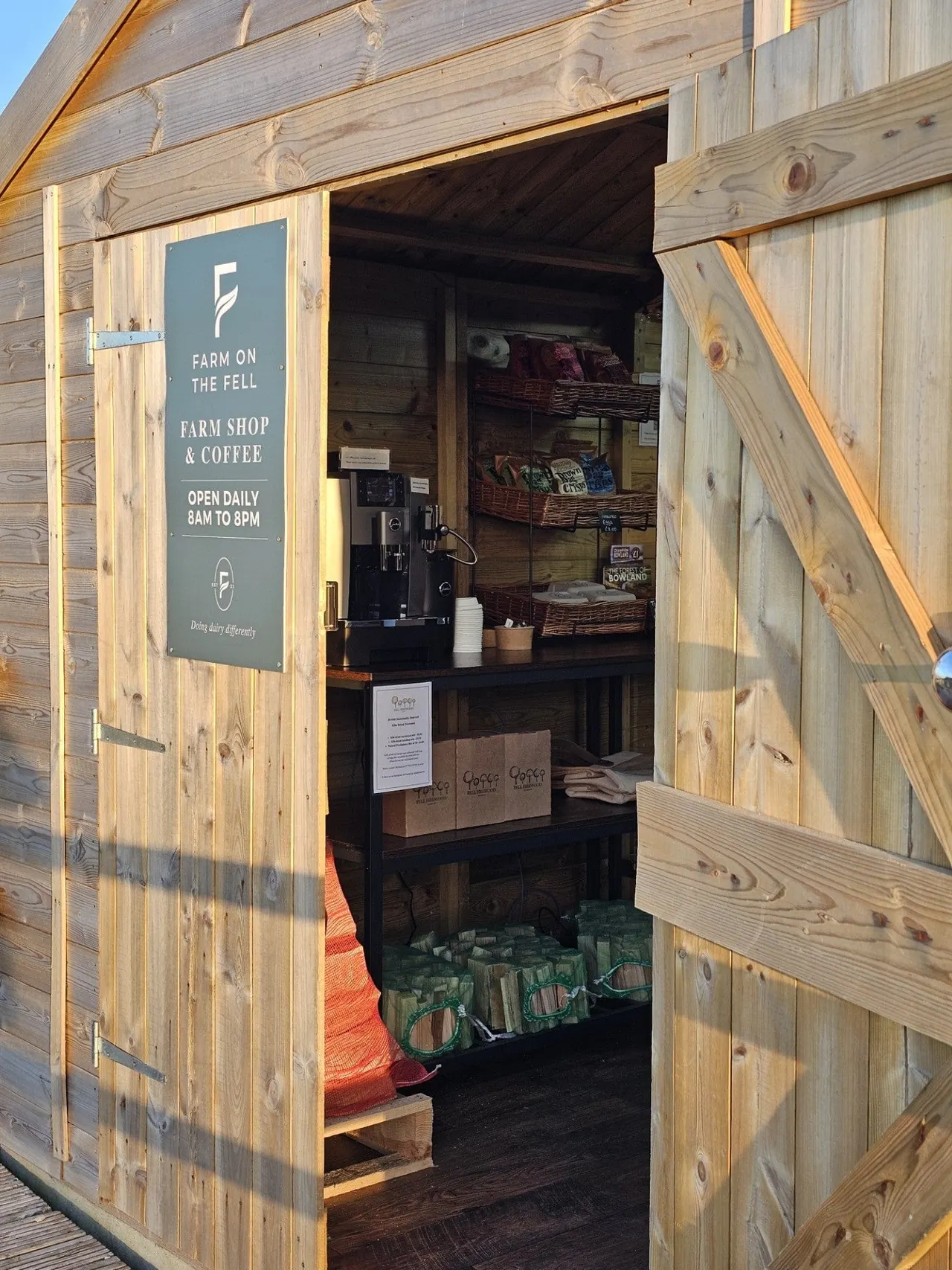 Wooden farm shop and coffee hut with open doors, displaying baskets of products and signage on a sunny day.