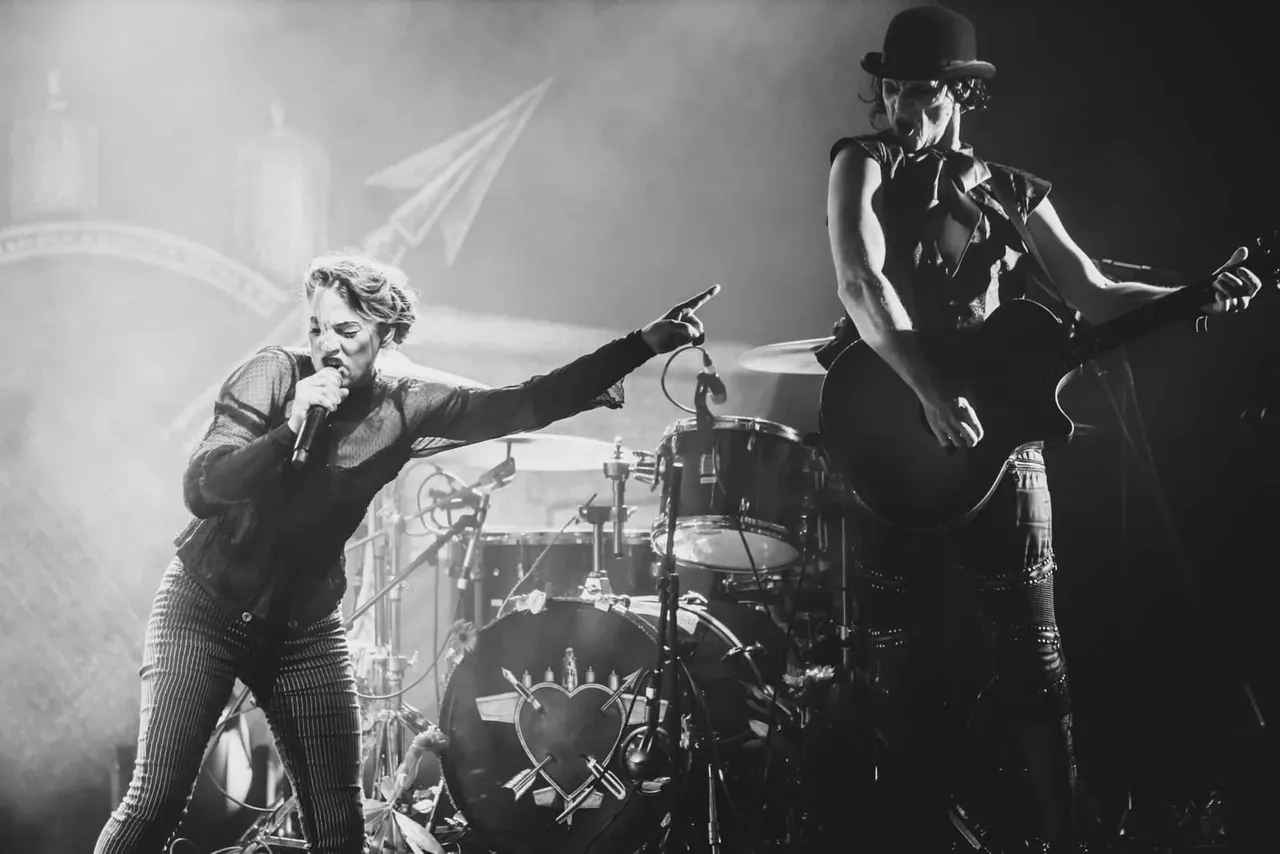 Two musicians performing on stage in black and white: vocalist with microphone on left, guitarist wearing bowler hat on right, with drum kit between them.