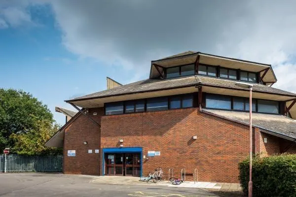 This is a PHOTOGRAPH.

Modern red-brick building with distinctive sloped roof features, blue entrance doors, and bicycles parked outside on a cloudy day.
