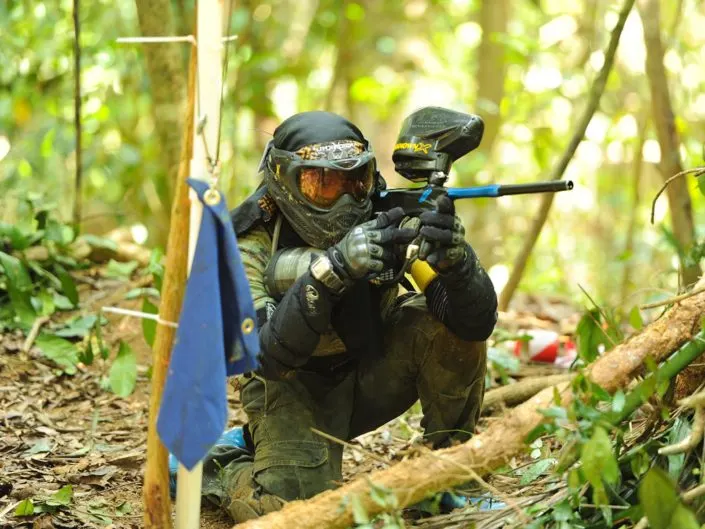 Player in protective gear aiming paintball marker in wooded course with fallen logs and vegetation.