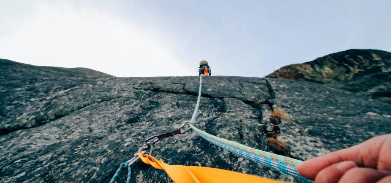 Person rock climbing on a cliff face with safety rope, viewed from below with another climber visible at the top.