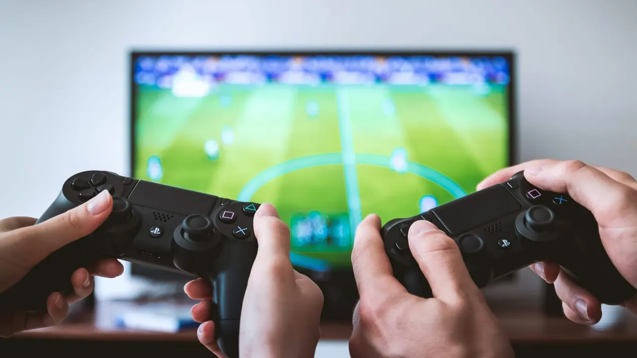 Two people holding gaming controllers whilst playing a football video game on a screen.