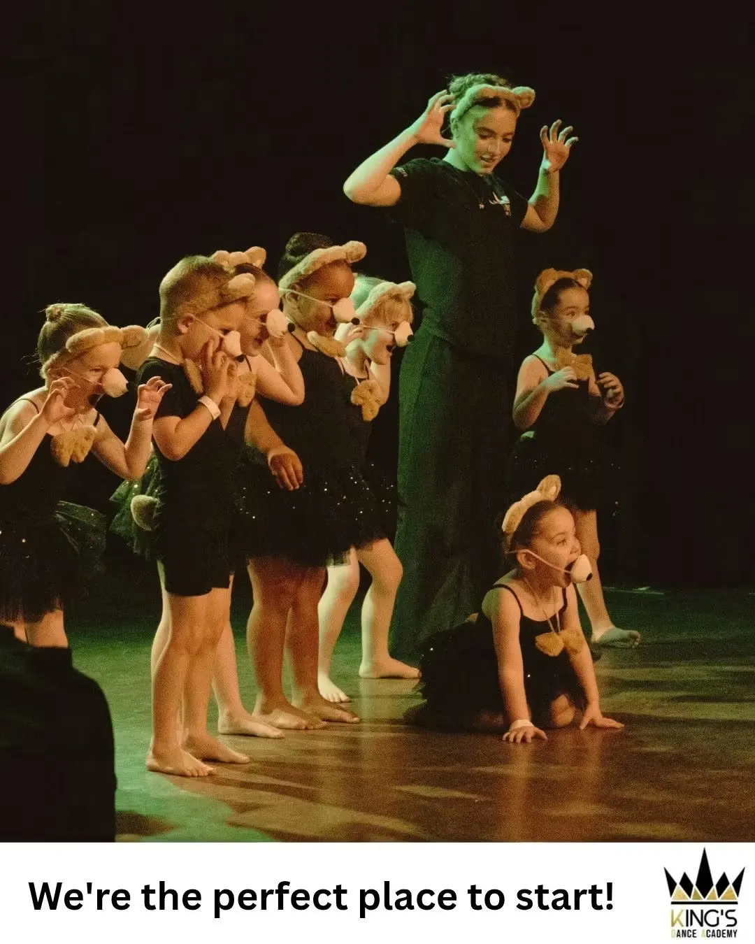 Children in bear costume ears and black outfits perform a theatrical dance routine on a lit stage.