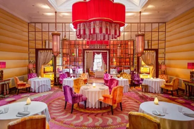 Elegant dining room with round tables, colourful chairs, and dramatic red chandelier suspended from coffered ceiling.