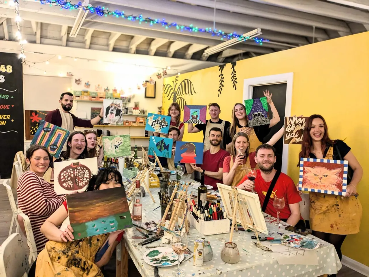 Group of adults holding finished artwork in a bright yellow studio space with paint-stained tables, art supplies, and blue festive decorations overhead.