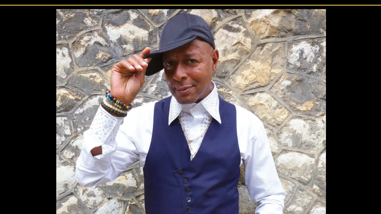 Man in blue waistcoat and cap tipping his hat against a stone wall backdrop.