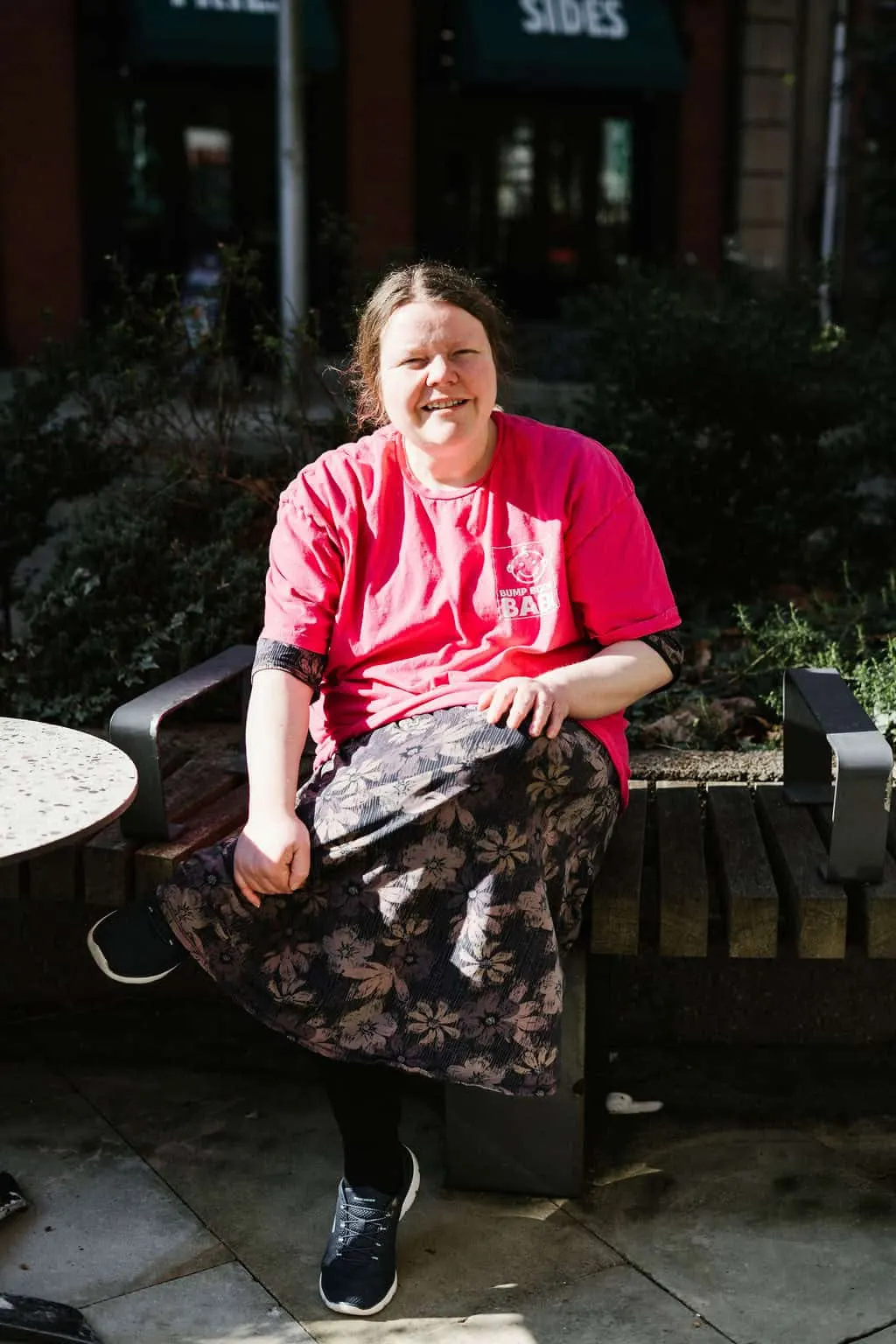 A smiling woman in a red t-shirt sits on a bench outside a venue entrance on a sunny day.
