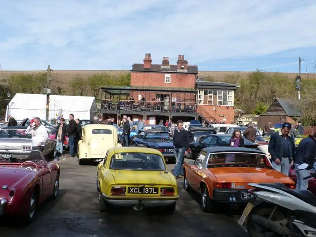Classic car enthusiasts gather at an outdoor venue with vintage vehicles on display and a brick building with terrace in the background.