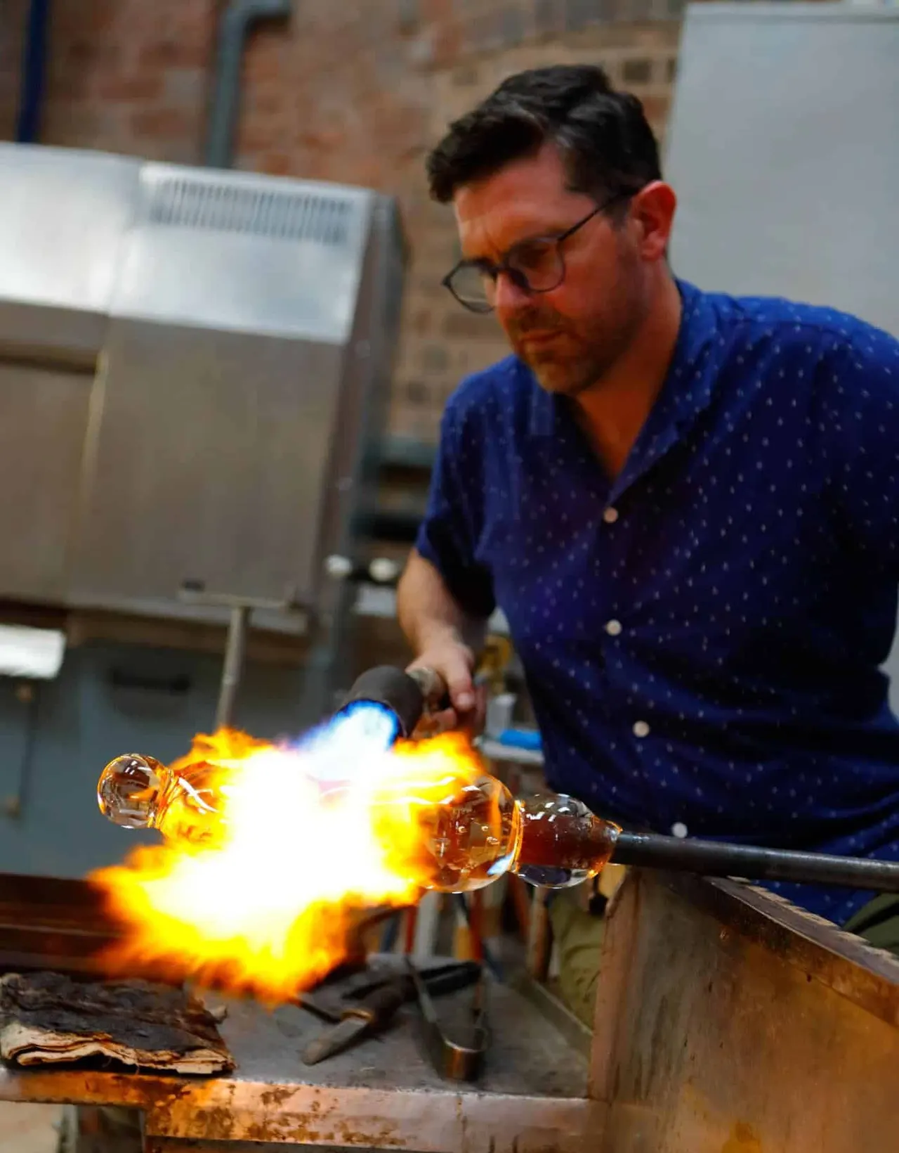 A craftsman uses a torch to heat molten glass whilst shaping it into decorative beads during a glassblowing demonstration.