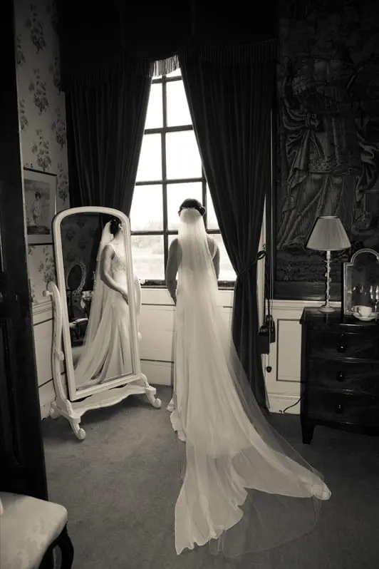 Bride in flowing white wedding gown and veil stands at tall window in ornate bedroom, viewing countryside through dark curtains.